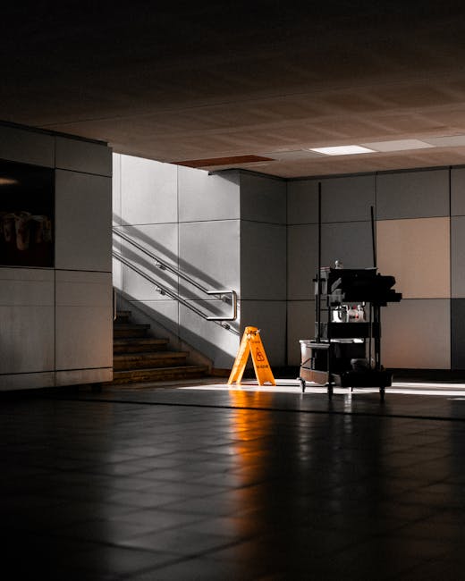 Interior of a commercial building showing a clean, tiled floor with a polished surface, illuminated by natural light from the right side. The scene includes a small cleaning cart with cleaning supplie